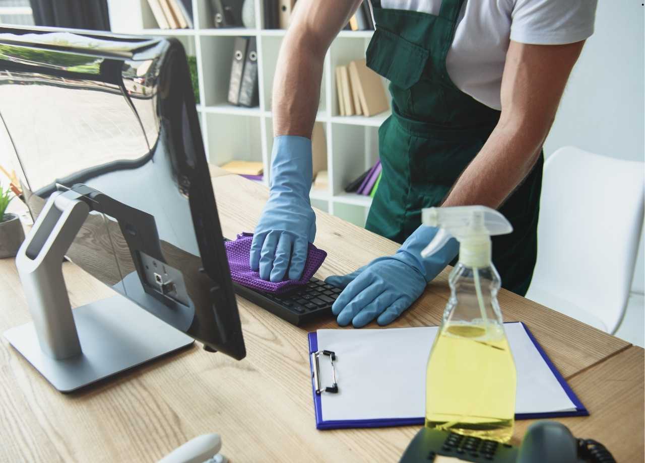 A man with gloves is cleaning the office desk where the computer is