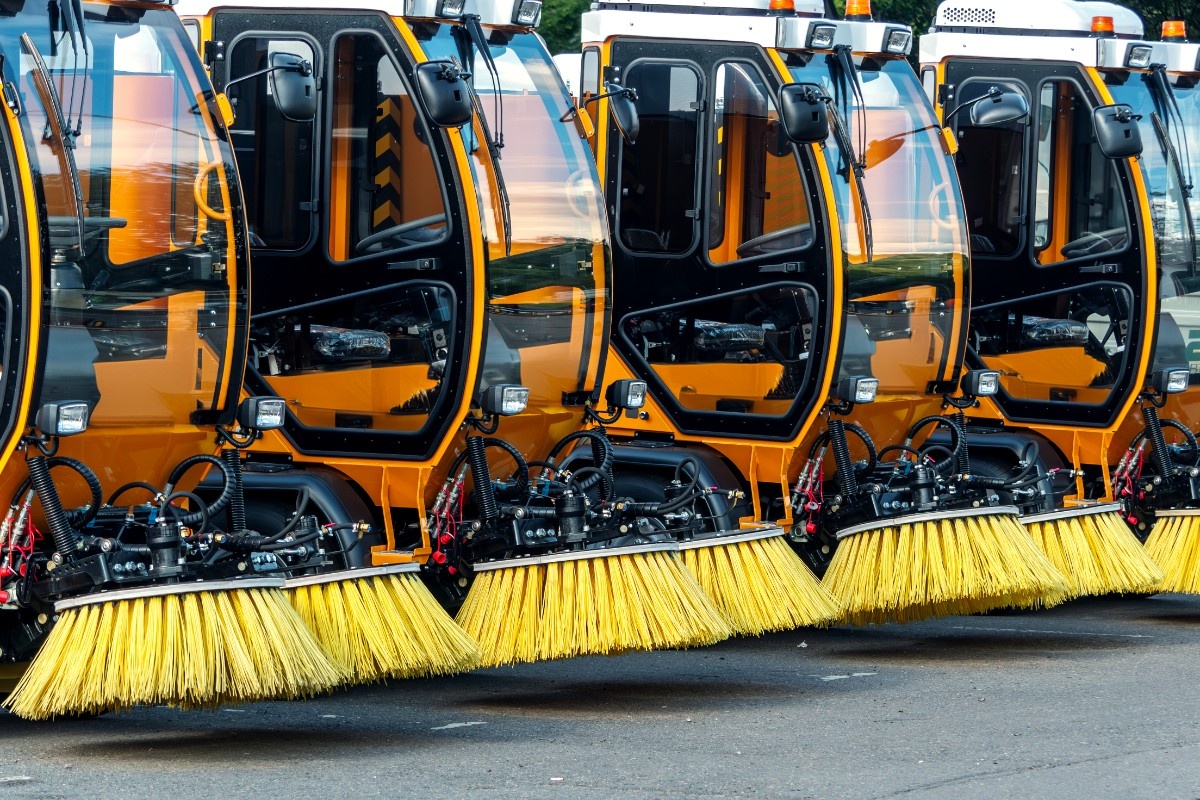Several orange street sweepers aligned in a row, with large brushes visible in the foreground