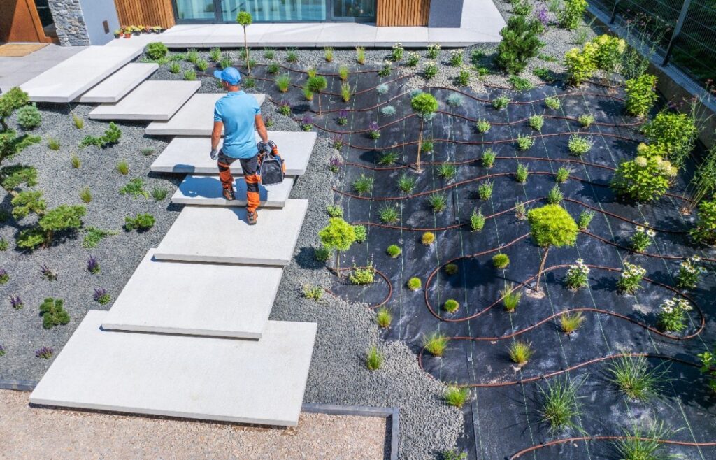 Worker Carrying Tools on Modern Walkway Through Landscaped Garden in Sunny Weather