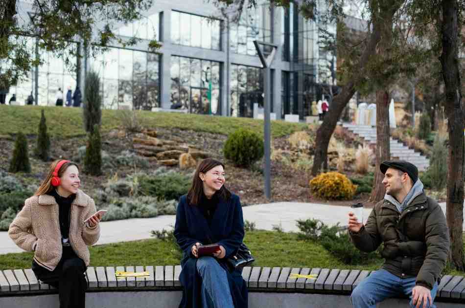 Three work colleagues sit outside their office building  enjoying landscape architecture behind them.