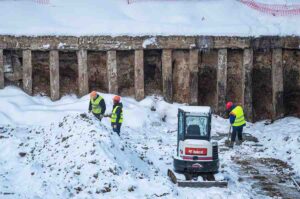 Three professionals wearing safety gear removing snow while snow remover is parked by the side