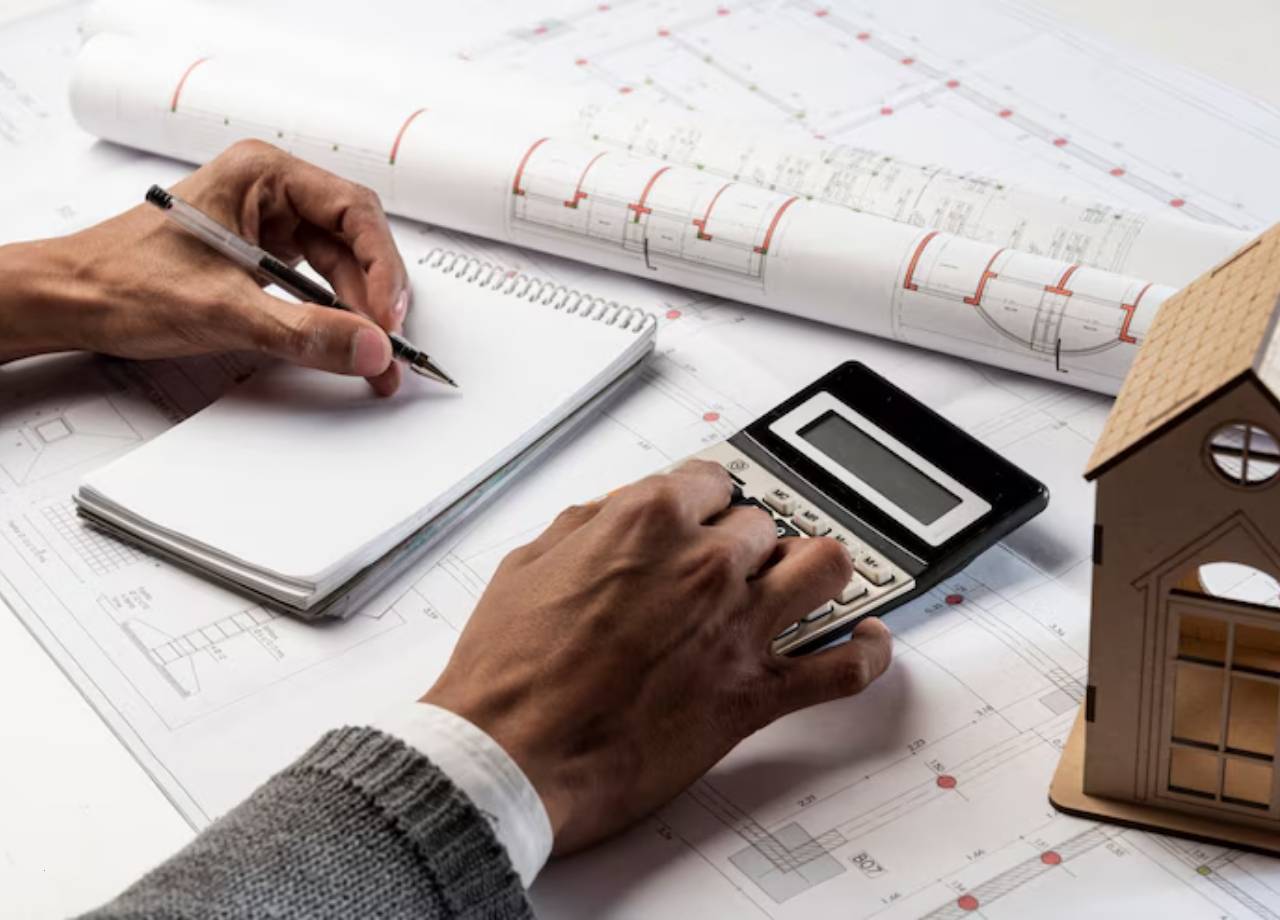 person with a calculator and notepad jotting down something with buidling plans on the desk