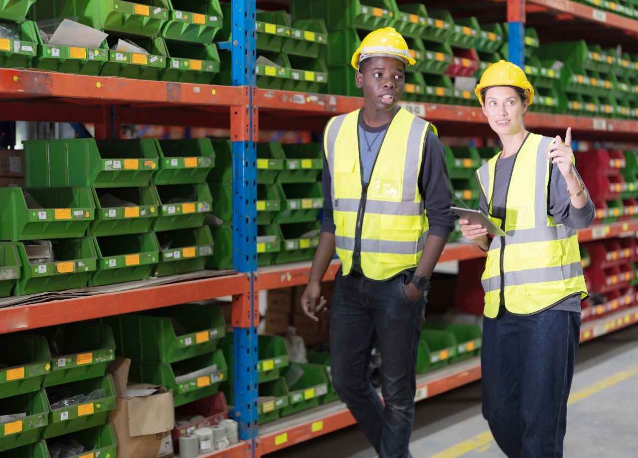 Two workers wearing safety gear, one female and one male. Walking in an inventory warehouse