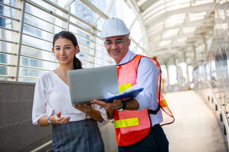 A man a nd woman both holding a laptop while man wears safety wear