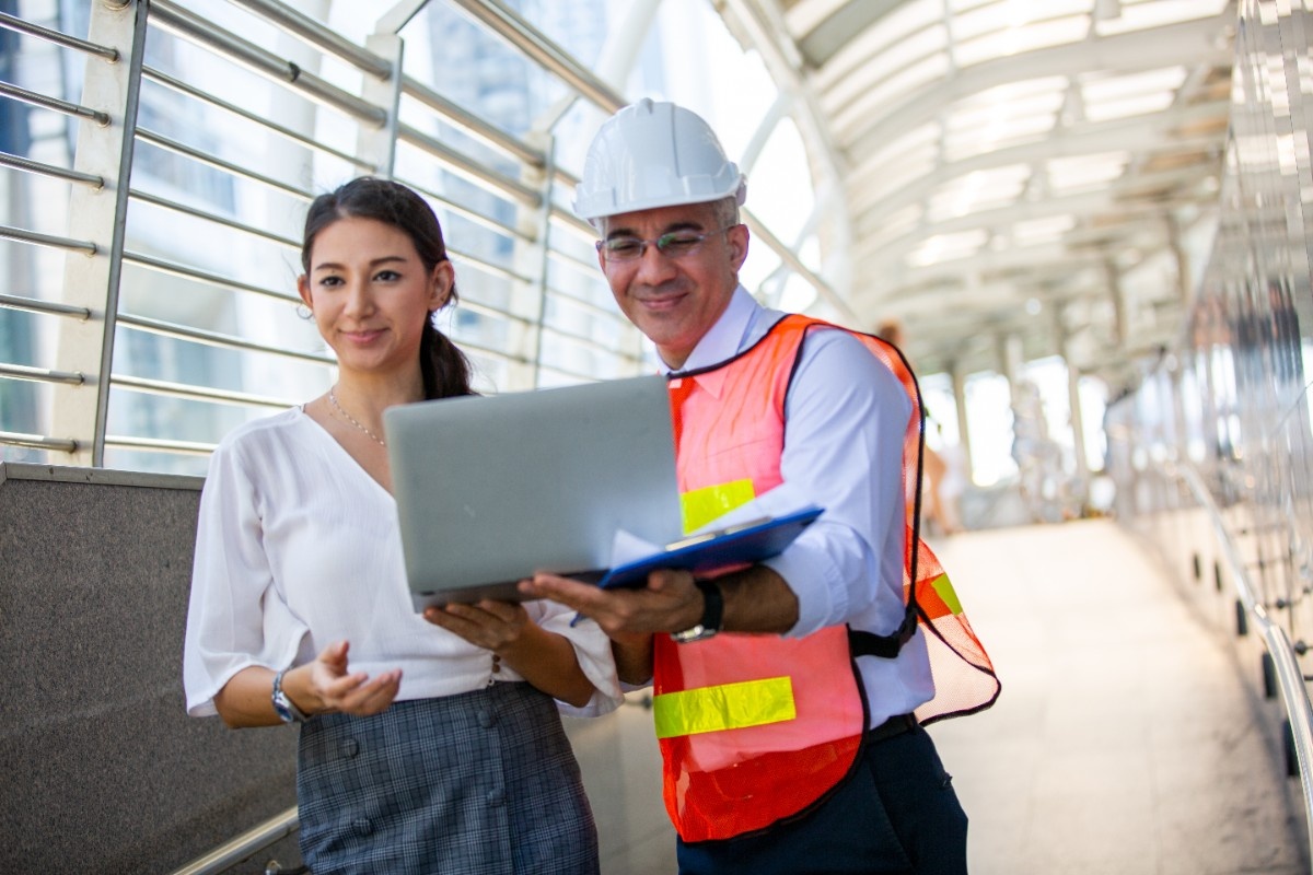 A man a nd woman both holding a laptop while man wears safety wear
