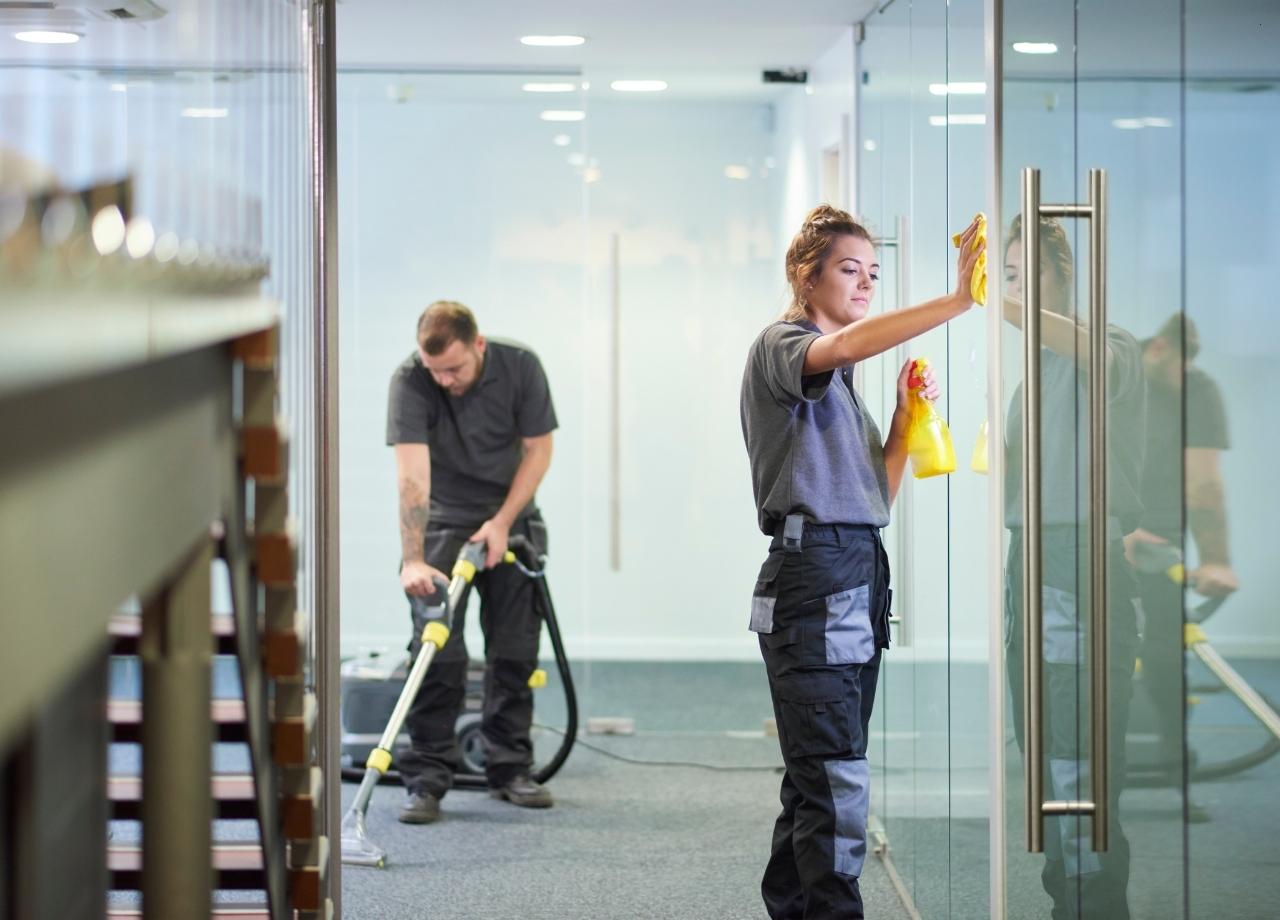 Professionals cleaning offices, woman cleaning windows while man vacuums floor