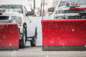 two white trucks with red snow plows sit side by side in a parking lot with snow falling