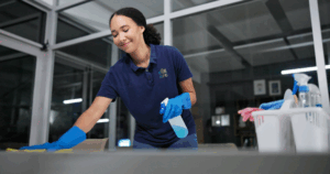 Woman in blue shirt uses spray bottle and rag to clean the surface of a table in a glassed-in office