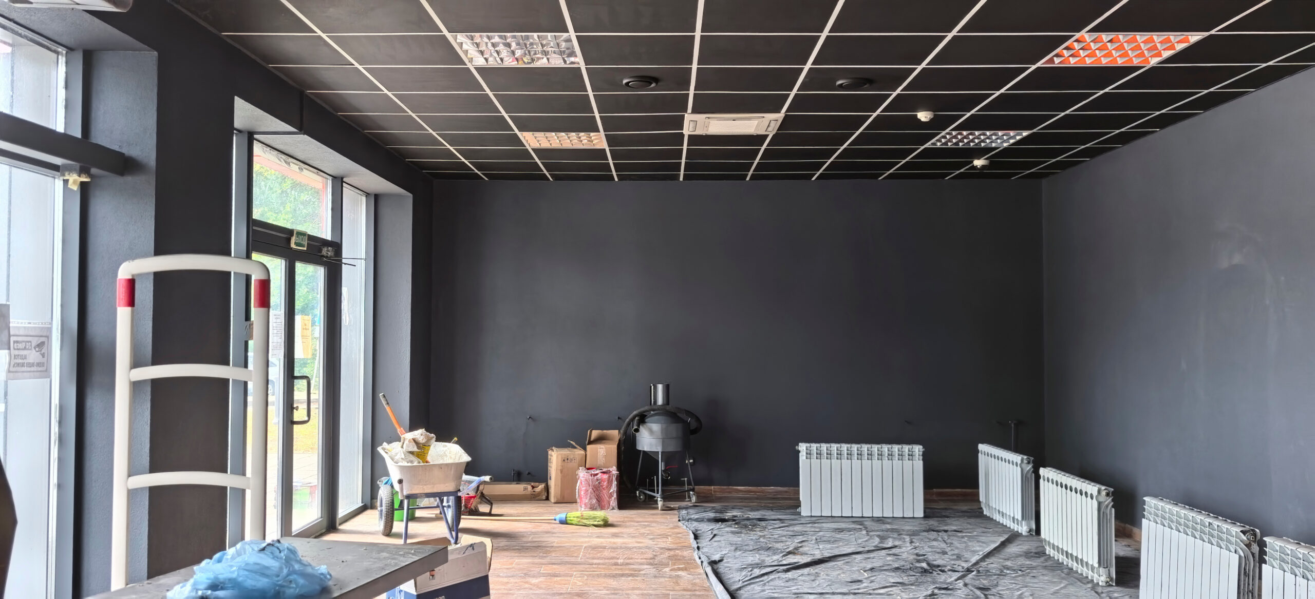 A vacant commercial space shows black walls and several radiators. Construction materials and tools are scattered around as the area is being prepared for future use.