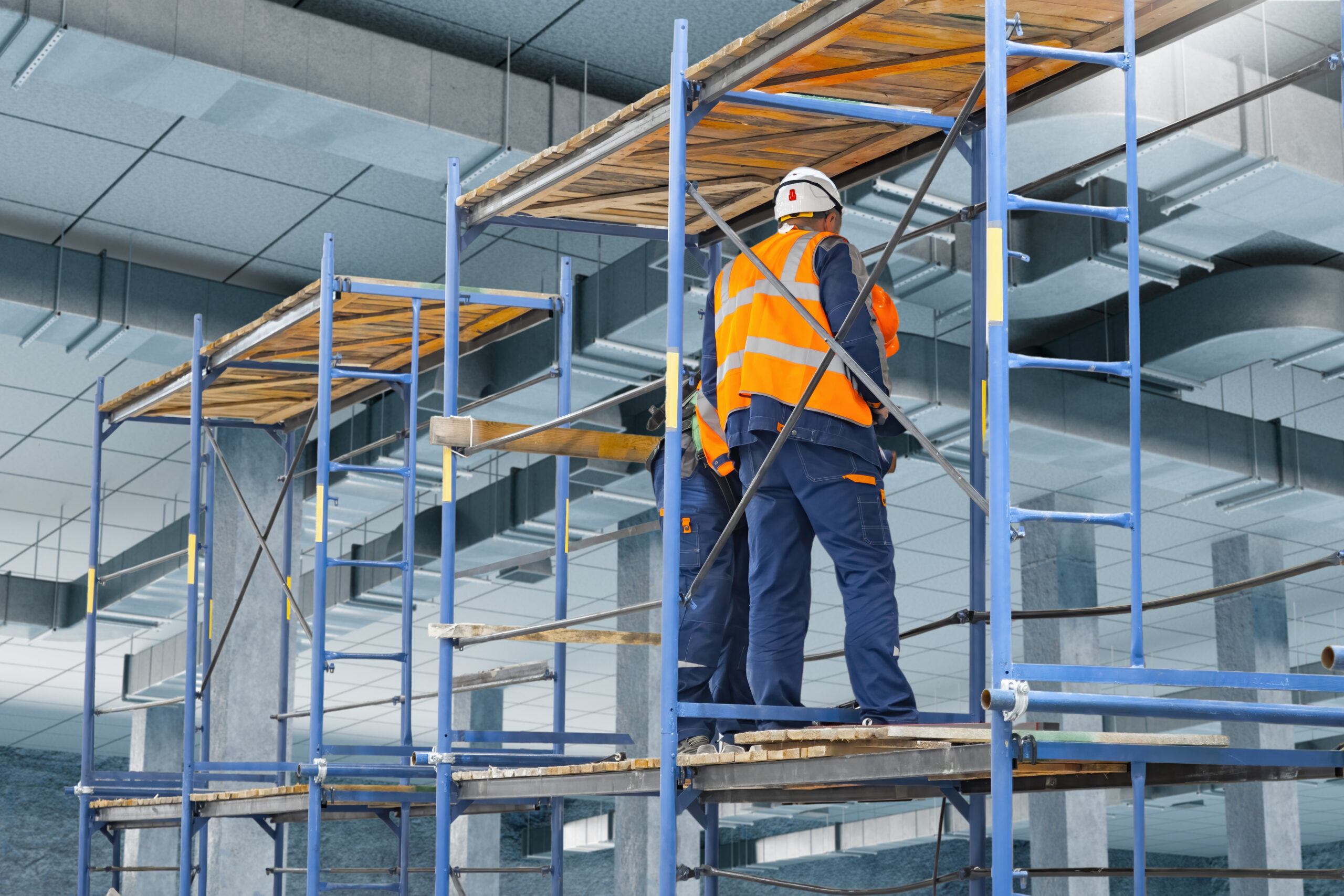 Construction site, Building renovation. Industrial workers in safety gear install ventilation systems using scaffolding inside commercial facility during interior construction works.