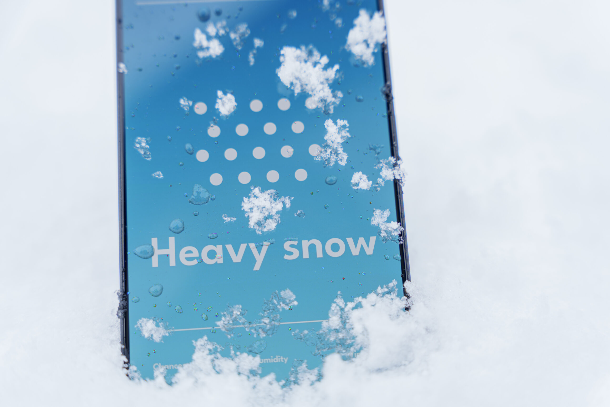 Smartphone standing in snow with heavy snow forecast on weather app screen, winter storm concept. High quality photo showing the High Cost of Waiting to Plan Your Commercial Snow Removal Program