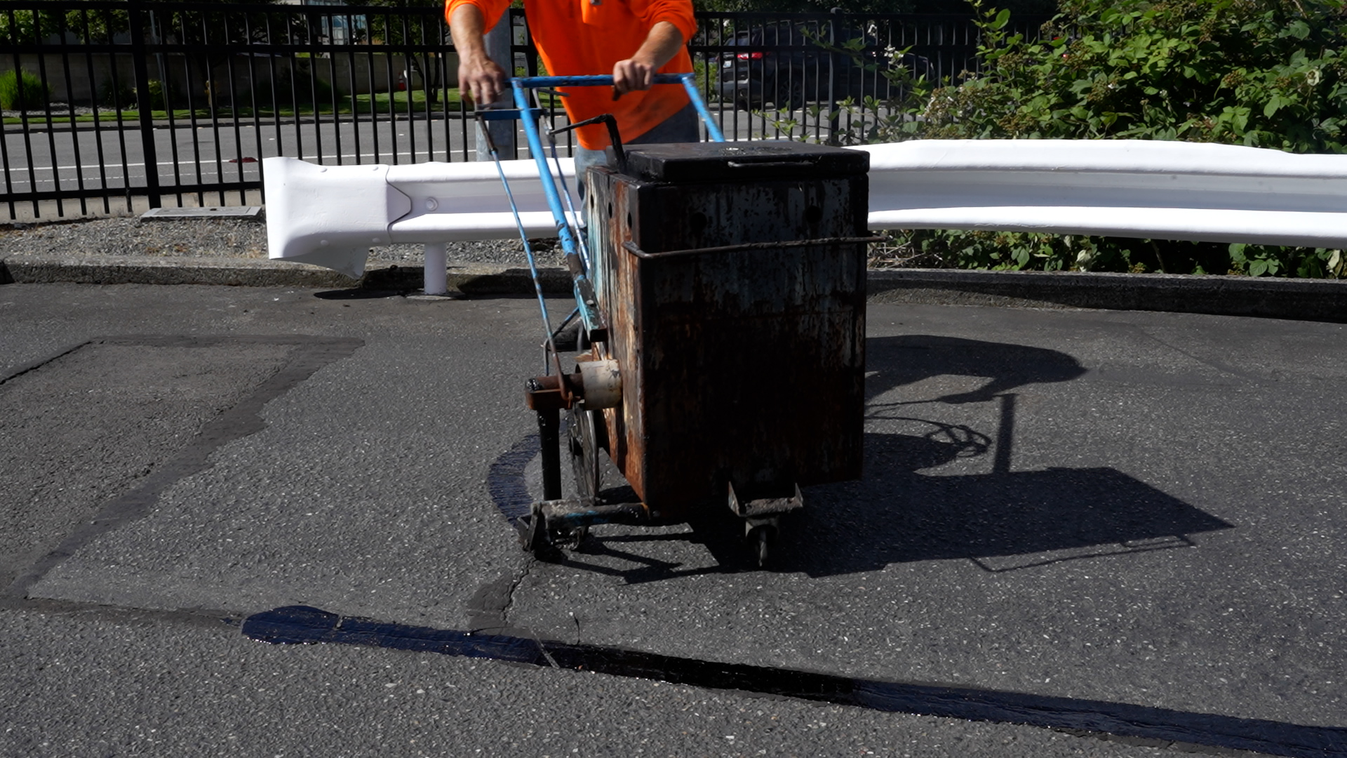 A worker in an orange shirt uses a crack-sealing machine to fill parking lot cracks
