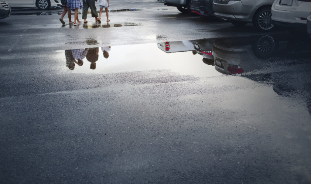 Wet parking spaces after hard rain fall with reflection of cars and walking people in puddles on the ground.