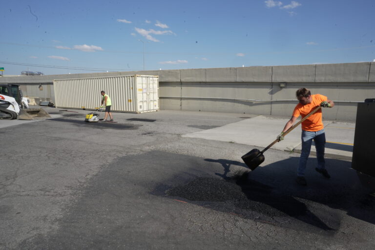 A crew performs September facility maintenance by filling potholes in a commercial parking lot with fresh asphalt.