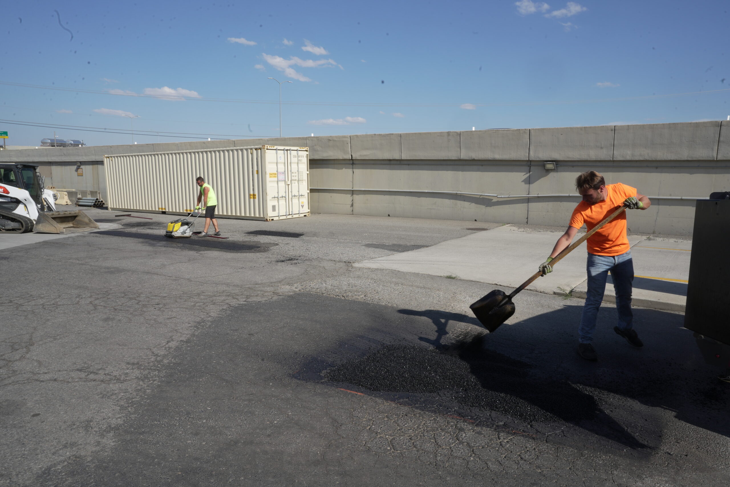 A crew performs September facility maintenance by filling potholes in a commercial parking lot with fresh asphalt.